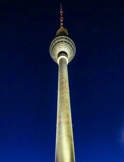 A low-angle nighttime photo of the Berlin TV Tower, its illuminated structure rising against a dark blue sky. The image is…