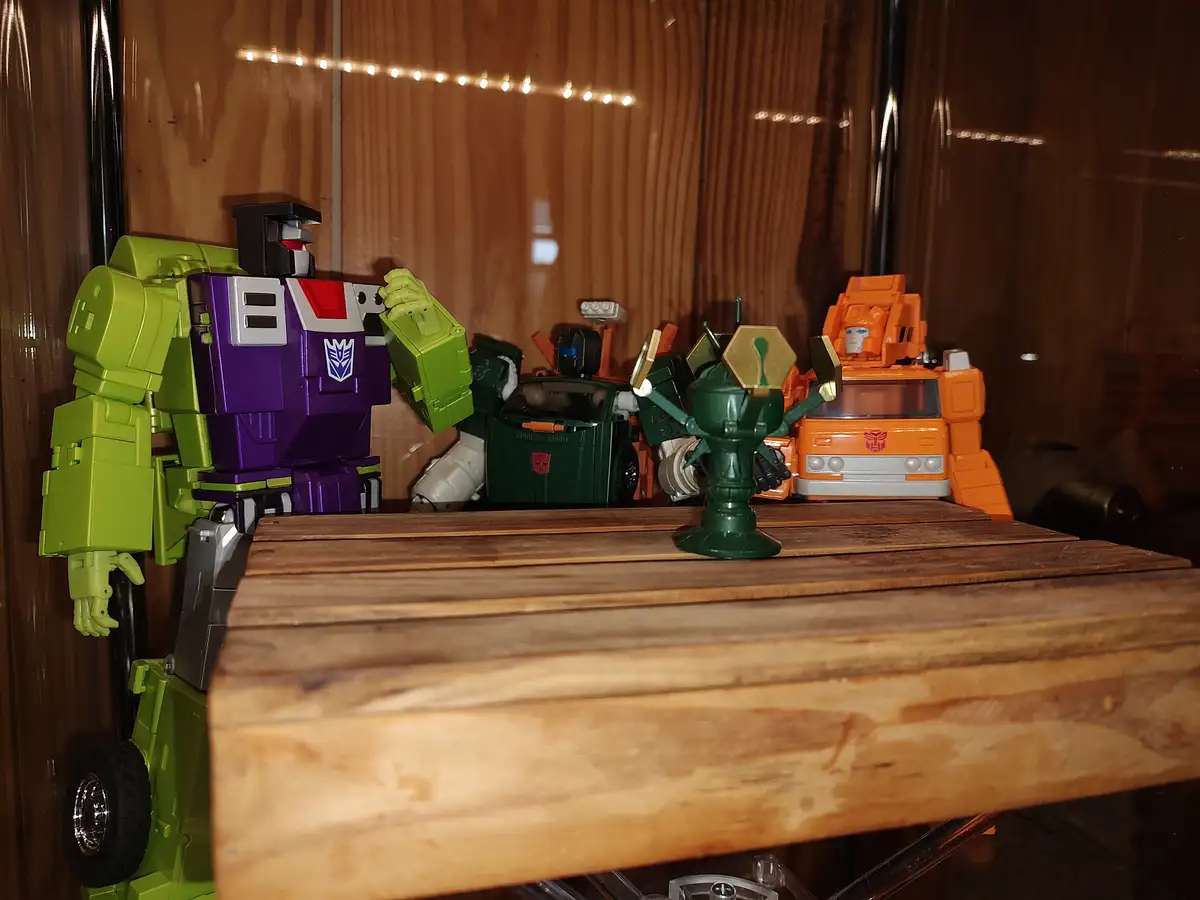 A green and purple Decepticon Transformer toy stands on a wooden shelf, gesturing toward a small green trophy-like object…