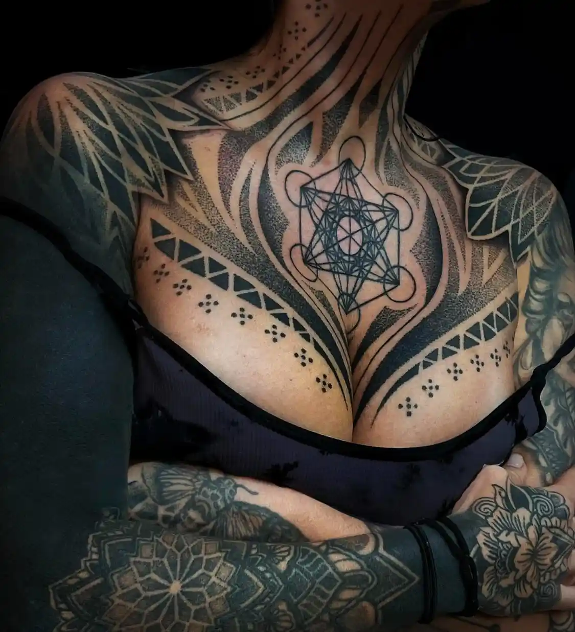 A person with extensive black ink tattoos covering their neck, shoulders, chest, and arms, wearing a dark top. The central…
