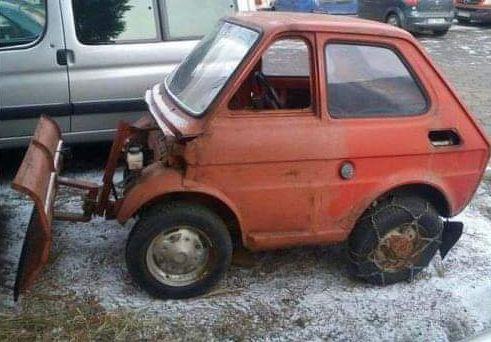 A rusted, orange, tiny car with a snow plow attachment bolted to its front, parked on snowy ground. The car's front end is…