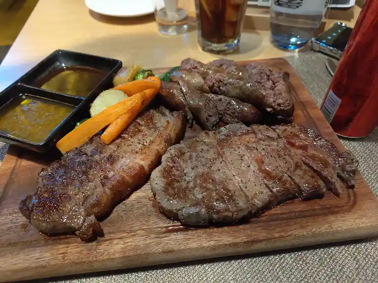 a wooden platter holds two sliced, seared steaks, a pile of sliced sausages, roasted carrots, potatoes, and broccoli…