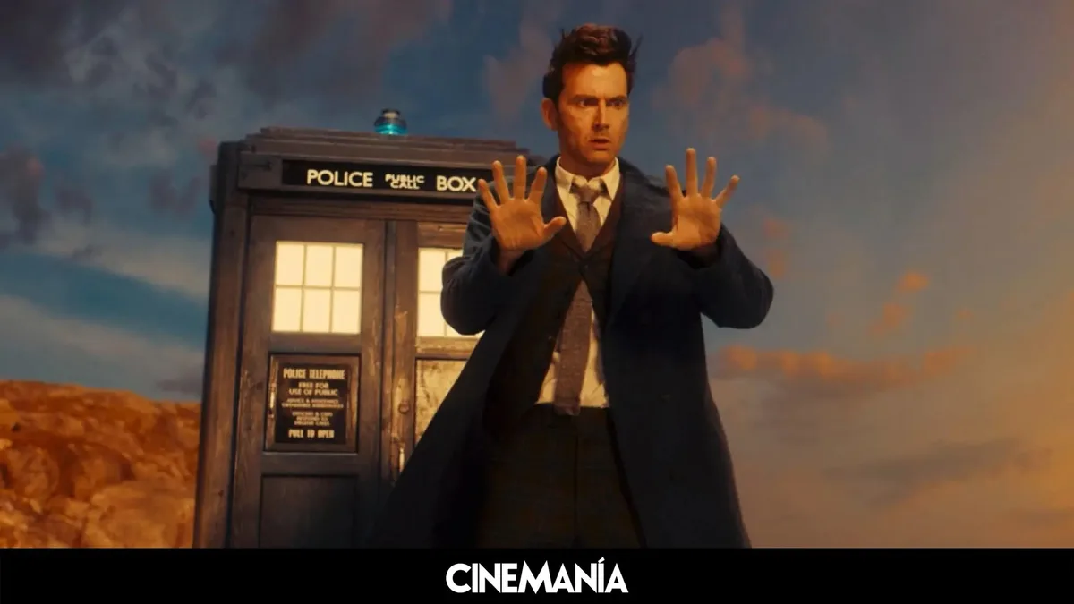 A man in a dark coat and tie stands in front of a blue police box with both hands raised, palms outward, in a scene from…