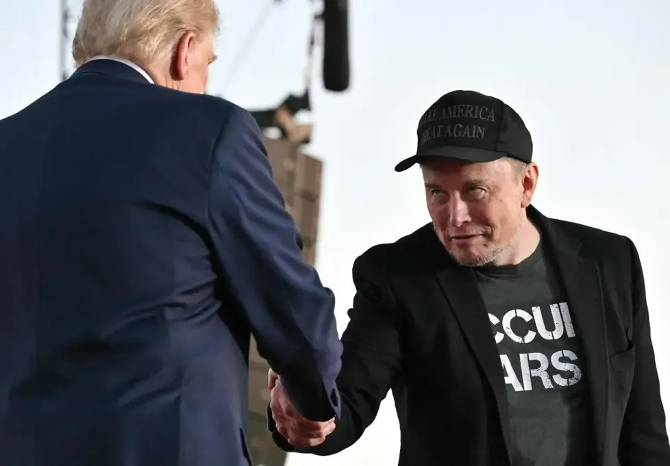 A man in a black cap with 'MAKE AMERICA GREAT AGAIN' text and a black t-shirt with 'CCUI ARS' partially visible shakes hands…