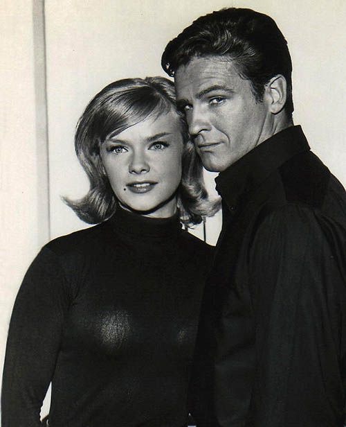 A black-and-white photo of a man and woman posing together, the man standing behind the woman with his arm around her. The…