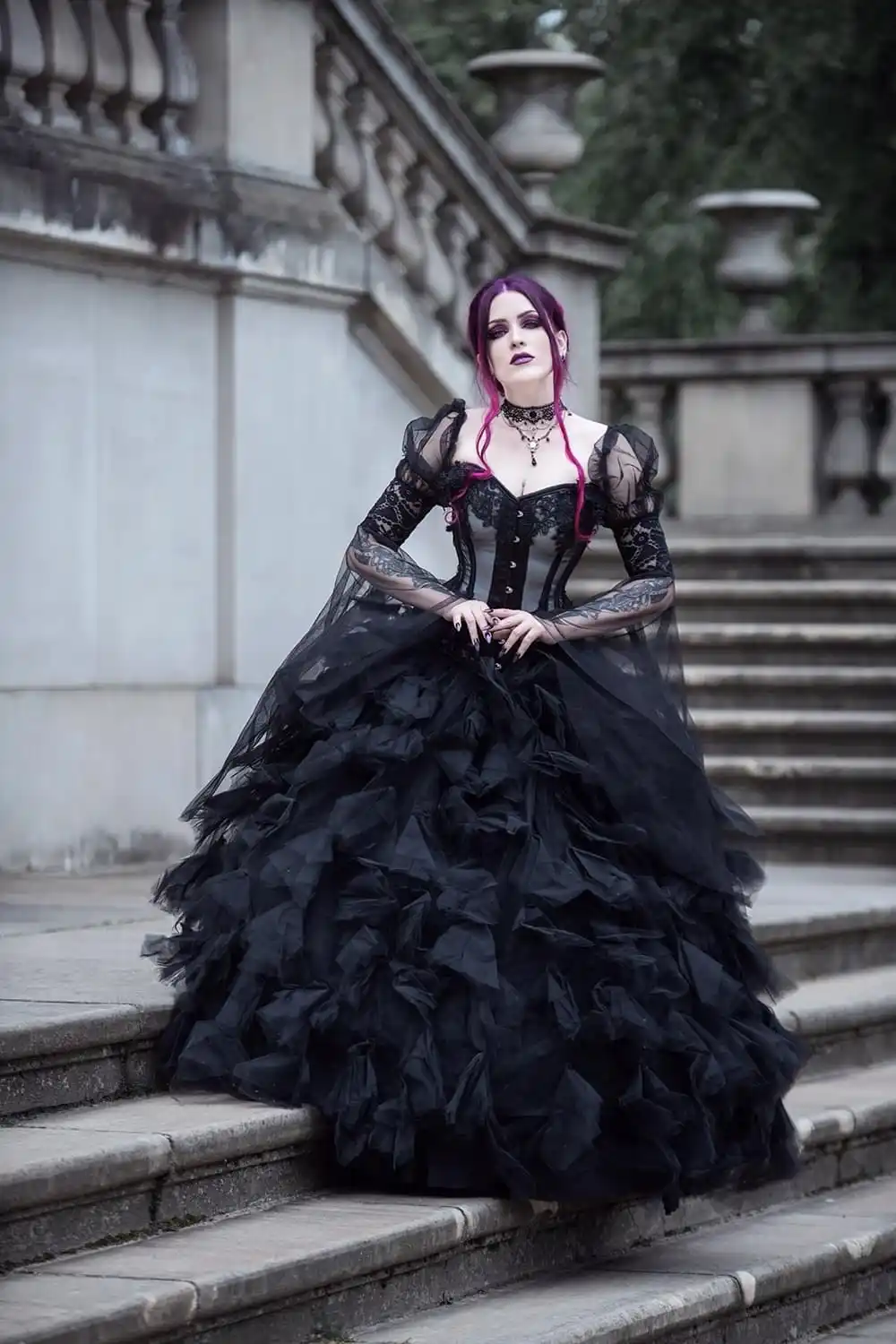 A woman with purple hair and gothic makeup sits on stone steps wearing an elaborate black gown with ruffles, lace, and…
