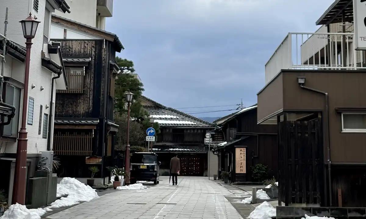 A person walks down a quiet, snow-dusted Japanese street lined with traditional wooden buildings and modern structures…