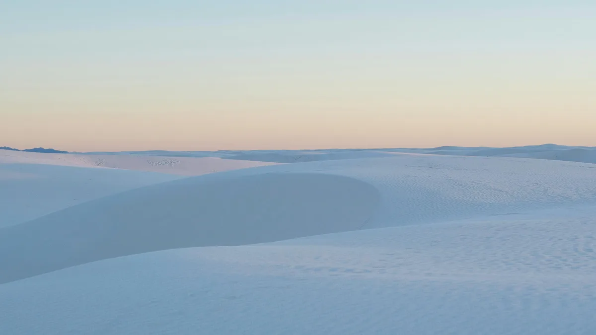 Soft pink and blue hues bleed into the dawn sky, casting a gentle glow on undulating white sand dunes, their delicate ridges…