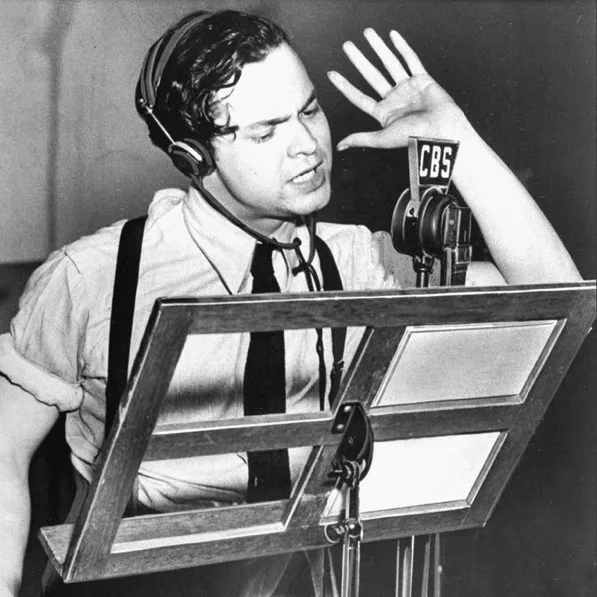 A black-and-white photo of a man in a studio wearing headphones and a tie, gesturing emphatically toward a CBS microphone…