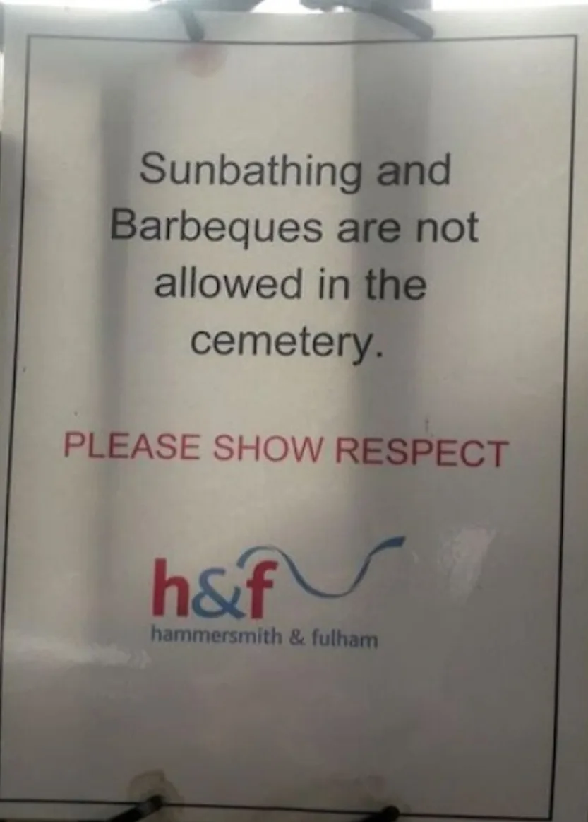 A white sign with black and red text forbids sunbathing and barbecues in a cemetery, urging visitors to 'PLEASE SHOW…