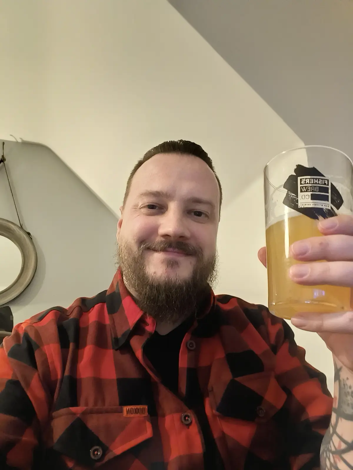 A man with a beard wearing a red and black plaid shirt holds up a glass of beer with a 'Fisher's Brew Co.' logo, smiling at…