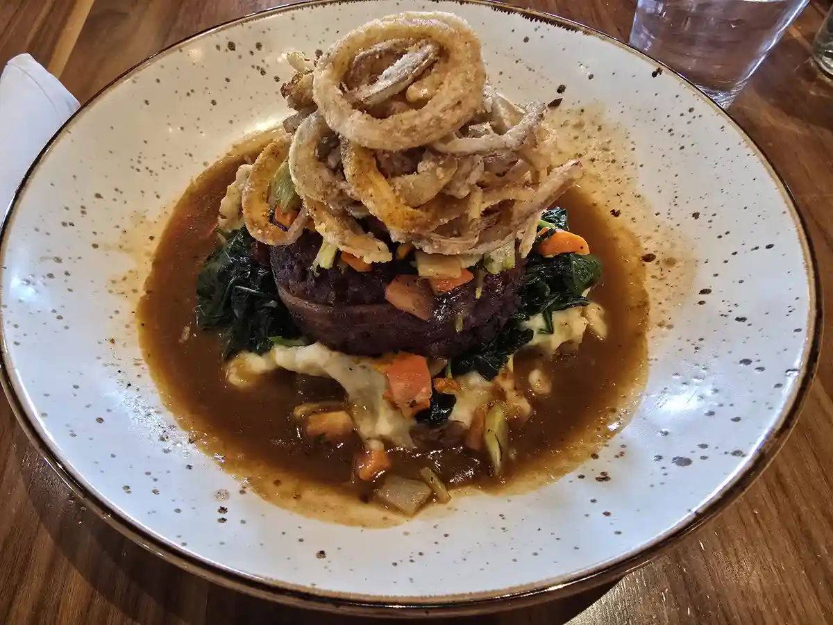 A close-up photo of a plated dish featuring a dark, round patty atop mashed potatoes, spinach, and carrots, all covered in a…