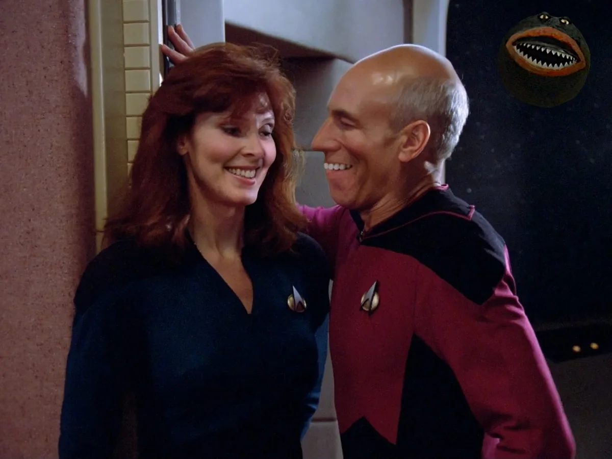 A man and woman in Star Trek uniforms smile at each other affectionately, with a cartoon frog head with sharp teeth floating…
