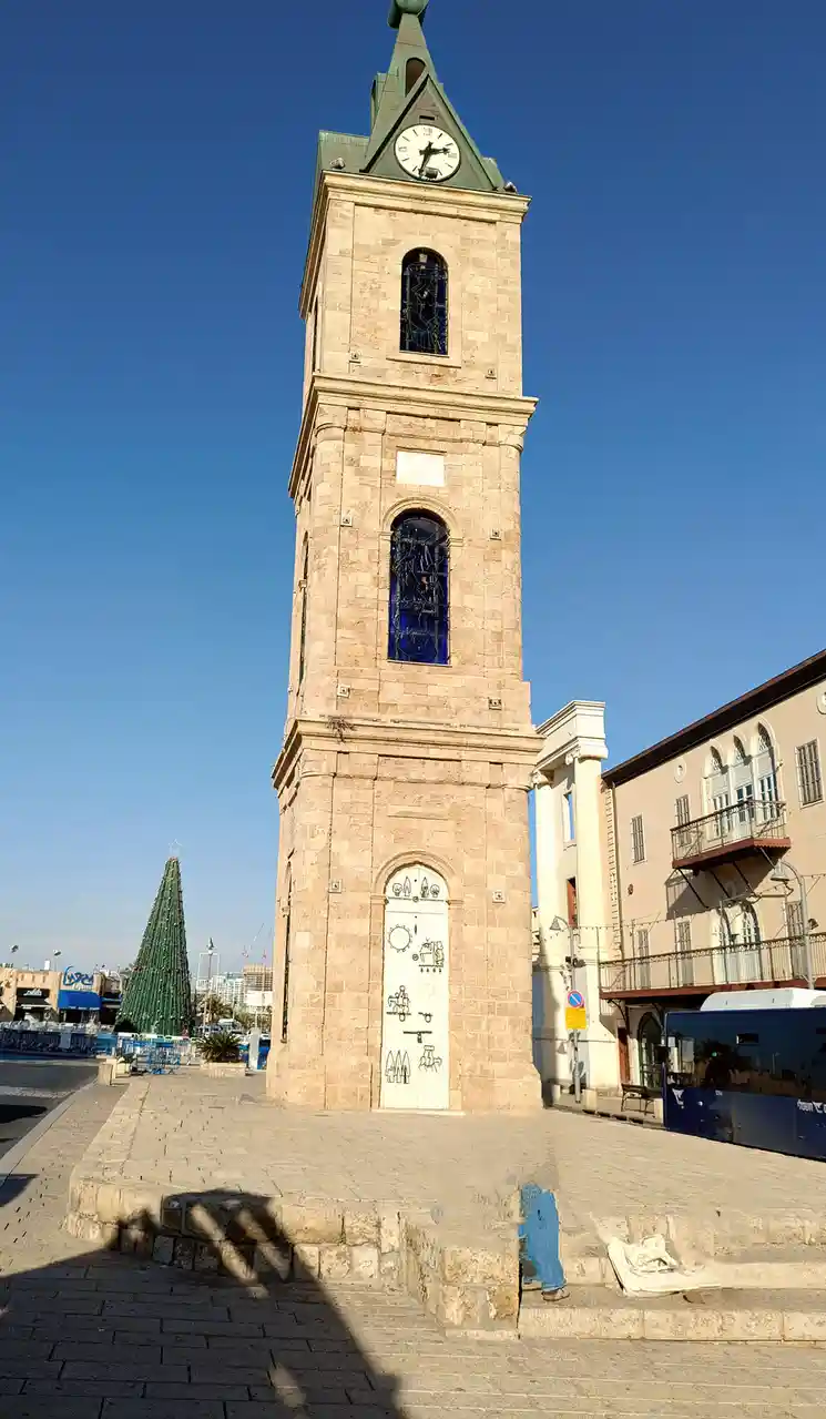 A tall stone clock tower with a green roof and clock face showing 3:00, set against a clear blue sky. A large green…
