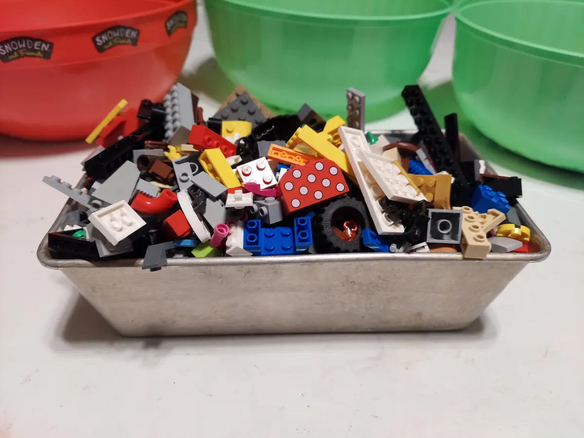 A metal baking pan is filled to overflowing with a chaotic mix of colorful Lego bricks and pieces of various shapes and…