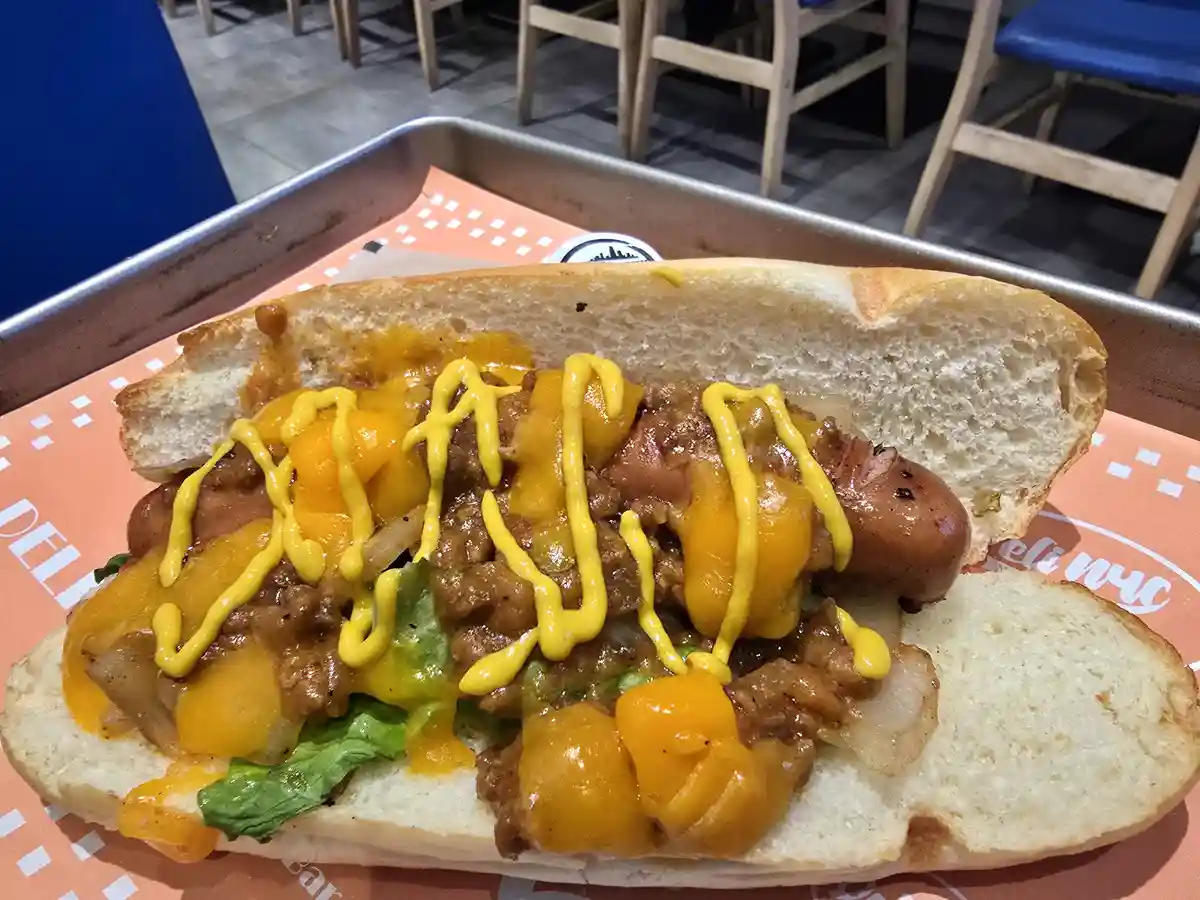 A close-up photo shows a hot dog in a bun overflowing with toppings including melted orange cheese, brown chili, yellow…
