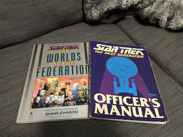 Two Star Trek FASA roleplaying game books are open on a gray couch, showing 'Star Trek: The Worlds of the Federation' and…
