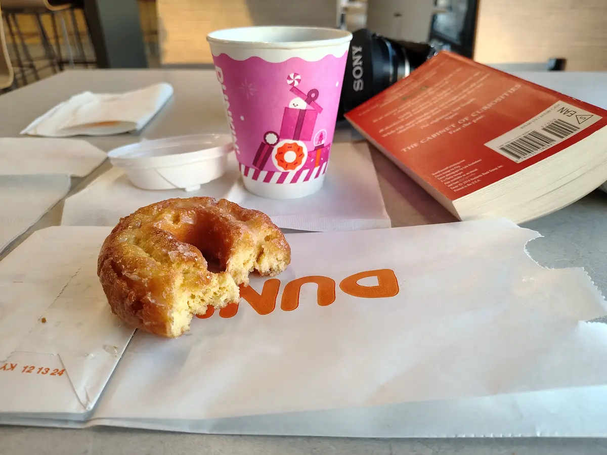 A partially eaten glazed donut sits on a Dunkin' paper bag next to a pink Dunkin' coffee cup, a book titled 'The Cabinet of…