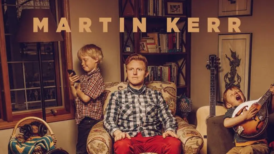 A man sits in a patterned armchair in a warmly lit living room, looking sternly at the camera. To his left, a young boy in a…