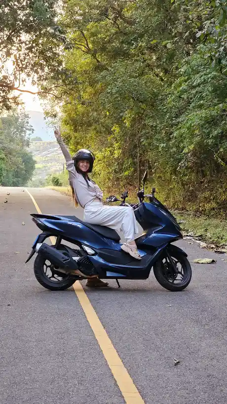A woman wearing a helmet and light-colored clothing sits on a blue scooter parked on a paved road lined with trees. She…