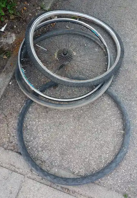Two bicycle wheels lie on a paved surface, one with a visible cassette and spokes, the other just a tire. The overlaid text…