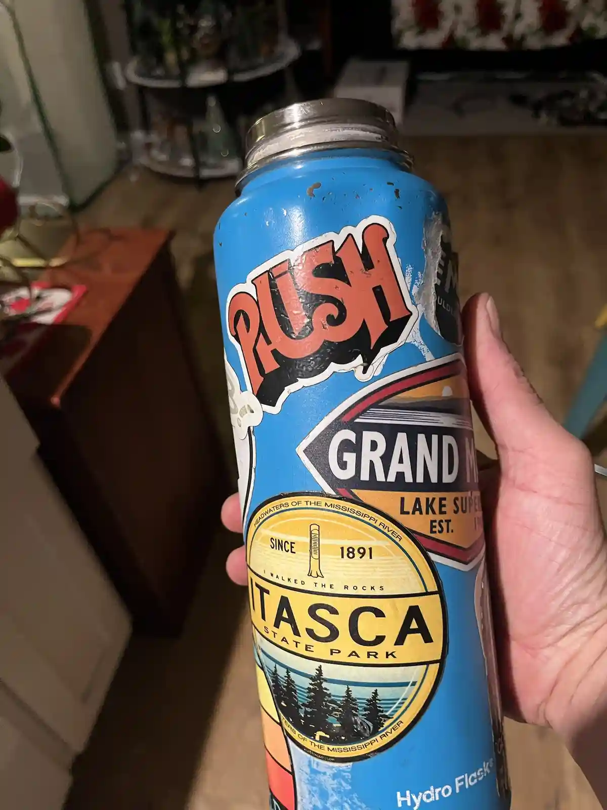 A hand holds a blue, weathered Hydro Flask water bottle with a vintage-style label featuring 'PUSH', 'GRAND MARSH', 'LAKE…