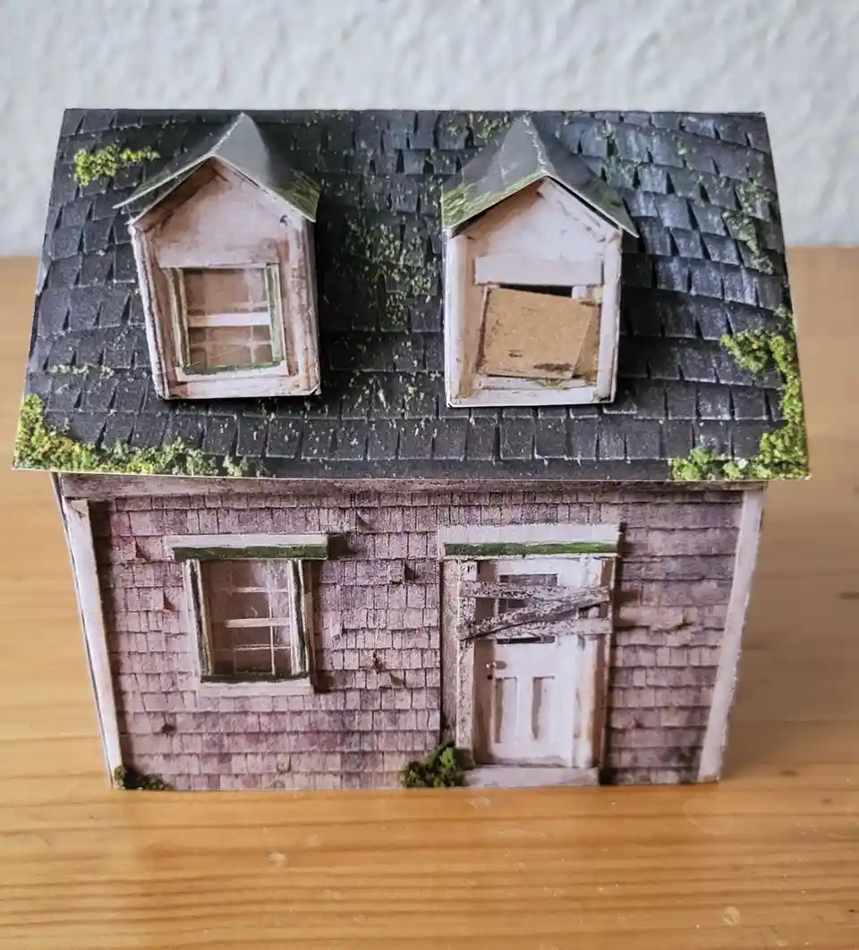 A handmade paper model of a dilapidated house with mossy shingles, boarded-up windows, and a broken door, sits on a wooden…