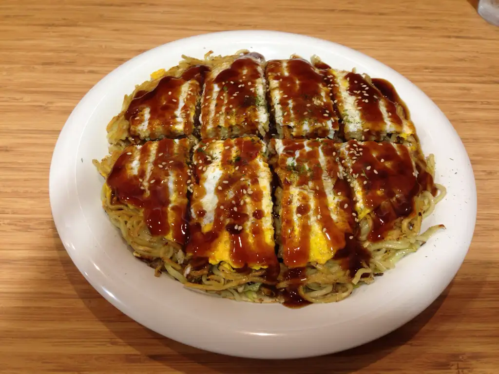 A round white plate holds a large, circular okonomiyaki dish, sliced into eight neat rectangular portions. The dish is…