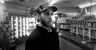 A black-and-white photo shows a man with a beard and cap standing in a store aisle, looking off to the side with a skeptical…