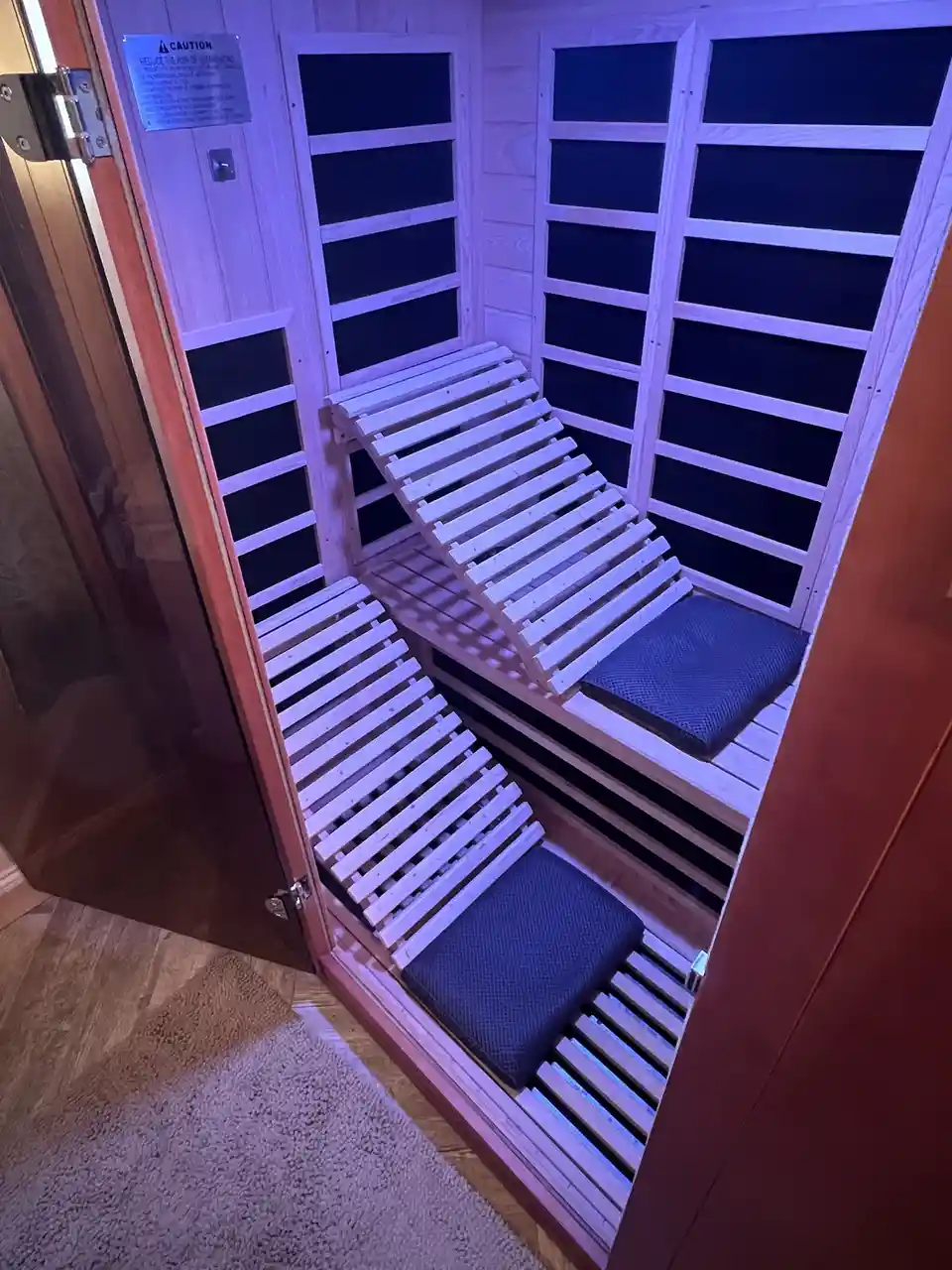 An empty wooden infrared sauna with tiered slatted benches and dark blue cushions, illuminated by purple ambient lighting. A…