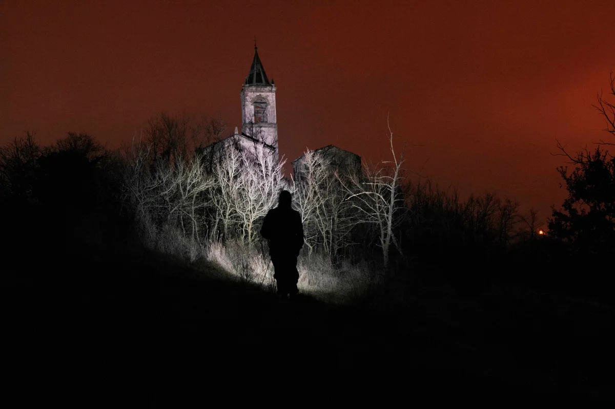 A person's silhouette stands in a dark field at night, illuminated by a flashlight beam, facing an old, eerie church with a…