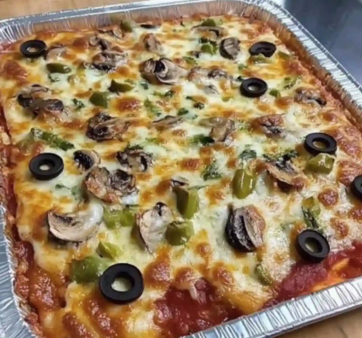 a square pizza in a foil pan with melted cheese, mushrooms, green peppers, and black olives, with text overlaid that reads…