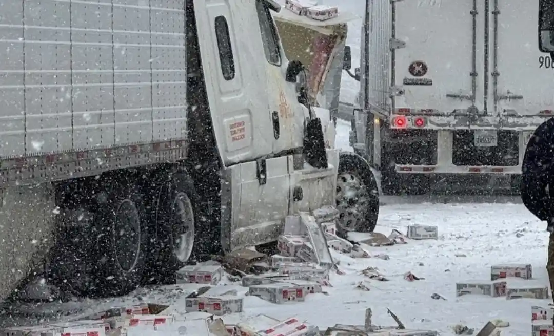 A white semi-truck is shown in a snowy scene with its cab severely damaged and crushed, spilling numerous cardboard boxes…