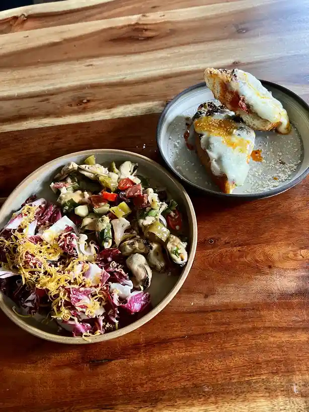 A top-down photo shows two plates of food on a wooden table: a bowl of salad with radicchio, cheese, and mussels, and a…