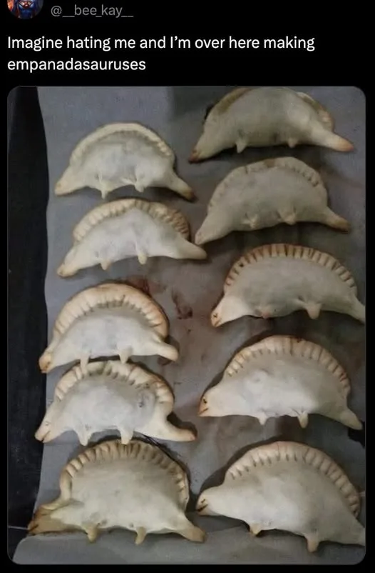 A baking sheet holds multiple empanadas shaped and arranged to resemble small, spiky dinosaurs, with the overlaid text…