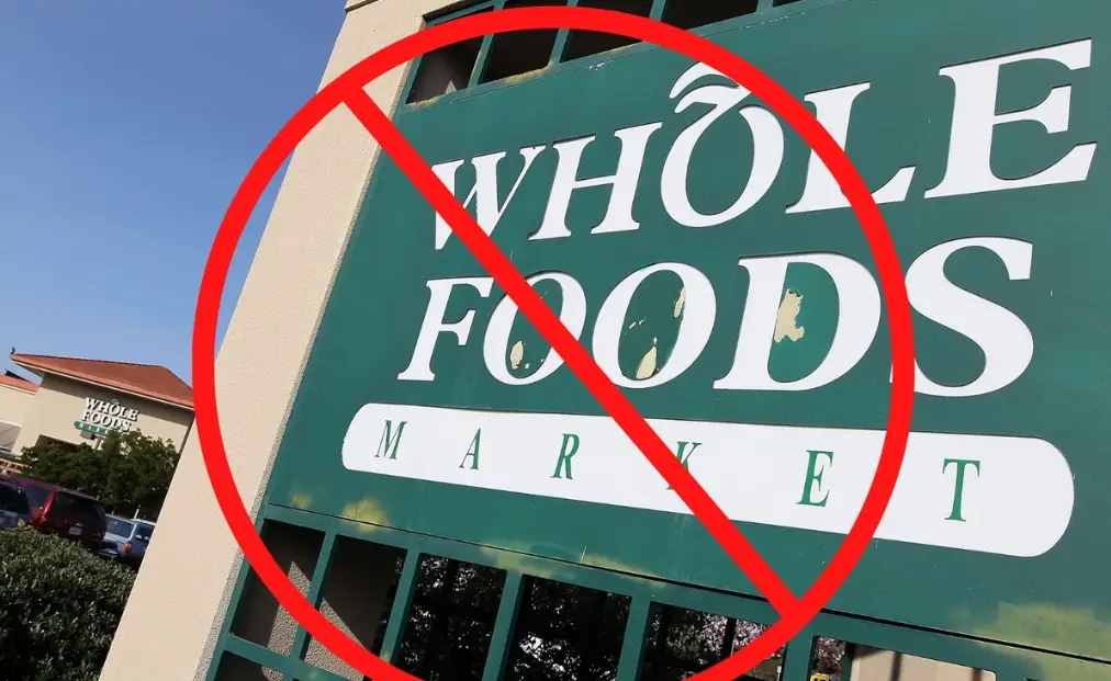 A green Whole Foods Market sign is overlaid with a large red circle and diagonal line, symbolizing prohibition or rejection…