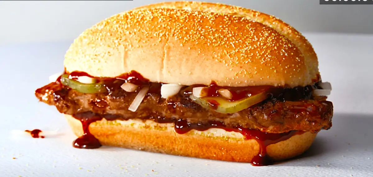 A close-up of a juicy burger with a sesame seed bun, dripping barbecue sauce, onions, and pickles, with the word 'selects'…
