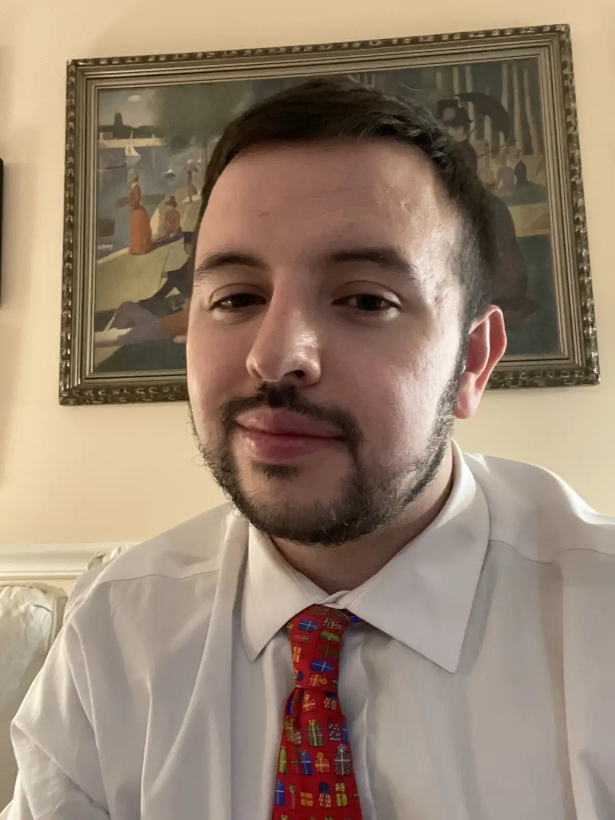 A man with a beard and mustache wearing a white dress shirt and a red tie covered in colorful gift boxes looks directly at…