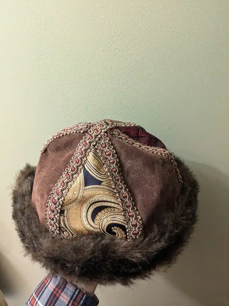A close-up photo shows the top of a brown, fur-trimmed hat with intricate gold and red embroidery, held against a plain…