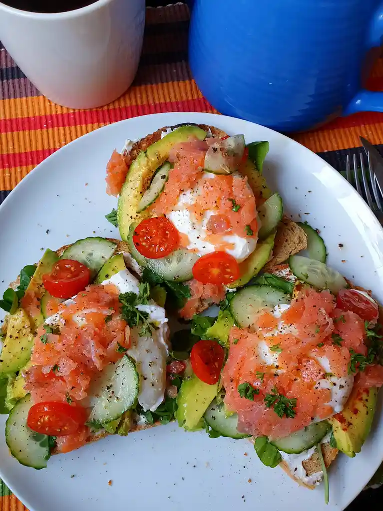 A white plate holds three open-faced sandwiches made with toasted bread, topped with avocado slices, cucumber rounds, cherry…