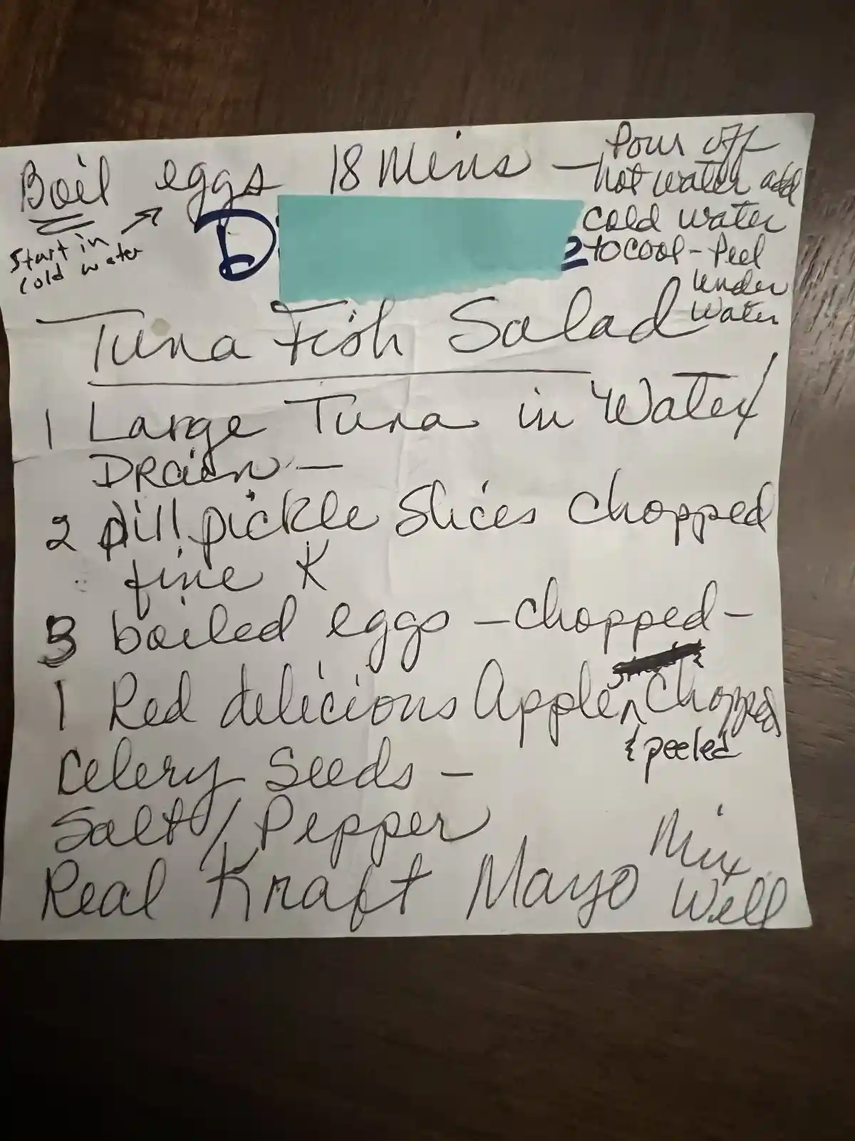 A handwritten recipe for 'Tuna Fish Salad' on a white paper, placed on a dark wooden surface. The recipe lists ingredients…