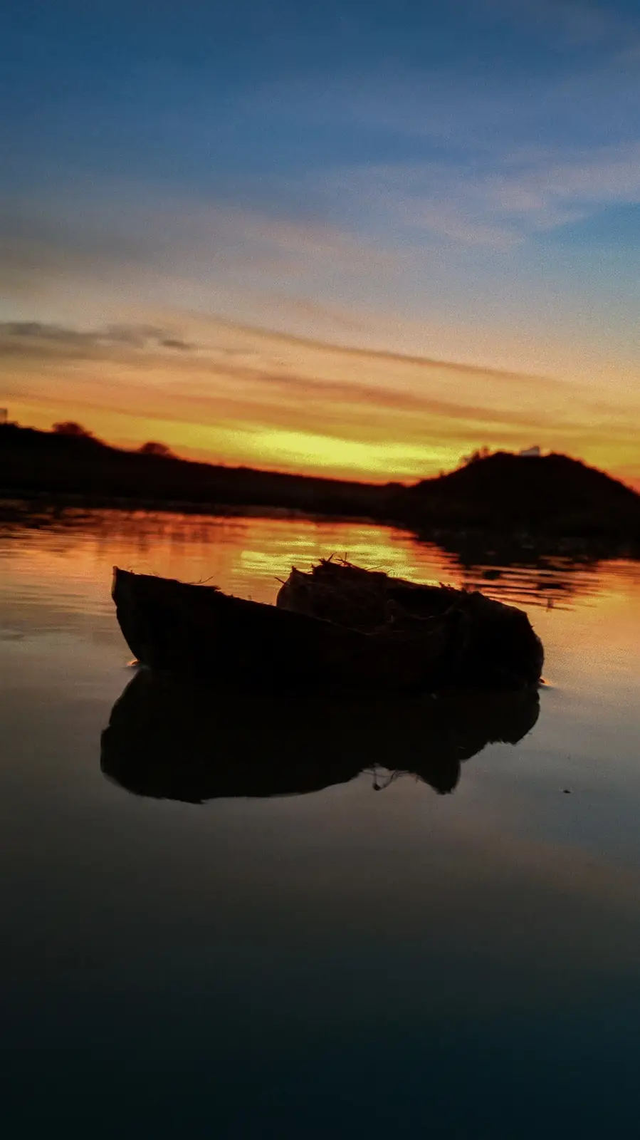 A serene sunset over water with a small, weathered boat floating in the foreground, its silhouette mirrored perfectly on the…