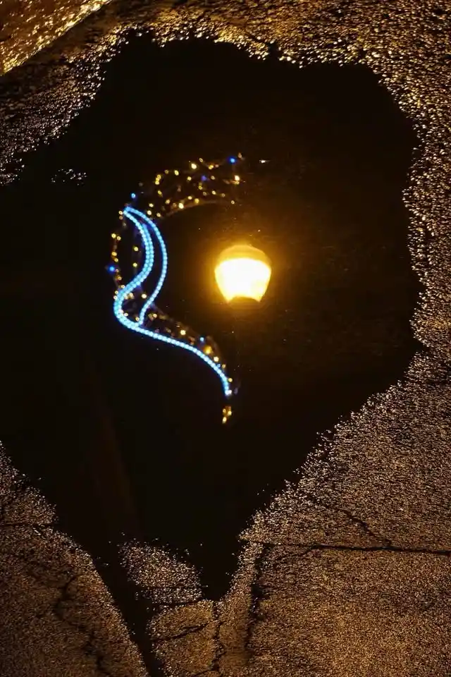 A nighttime photo shows a dark puddle on asphalt reflecting a streetlamp and a blue light trail, resembling a crying face…