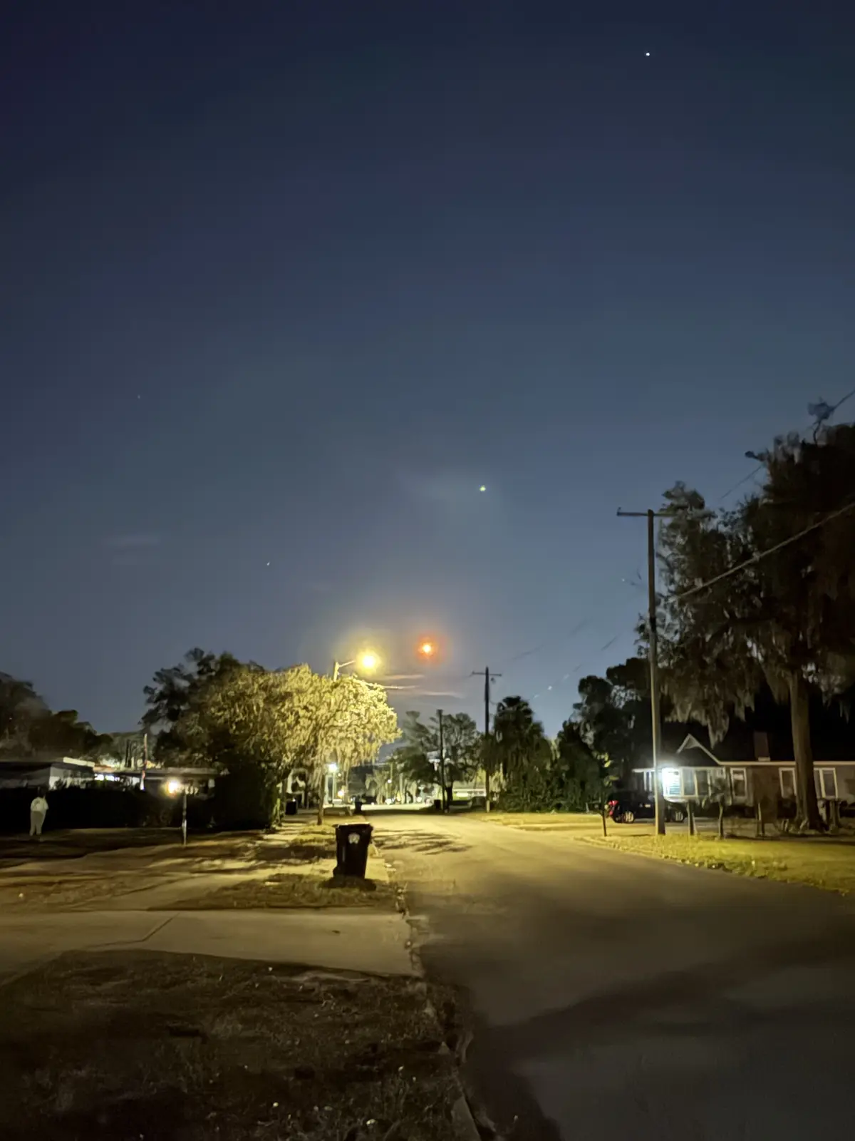 A nighttime street scene with streetlights, trees, houses, and a trash can, where a red dot in the sky is humorously…