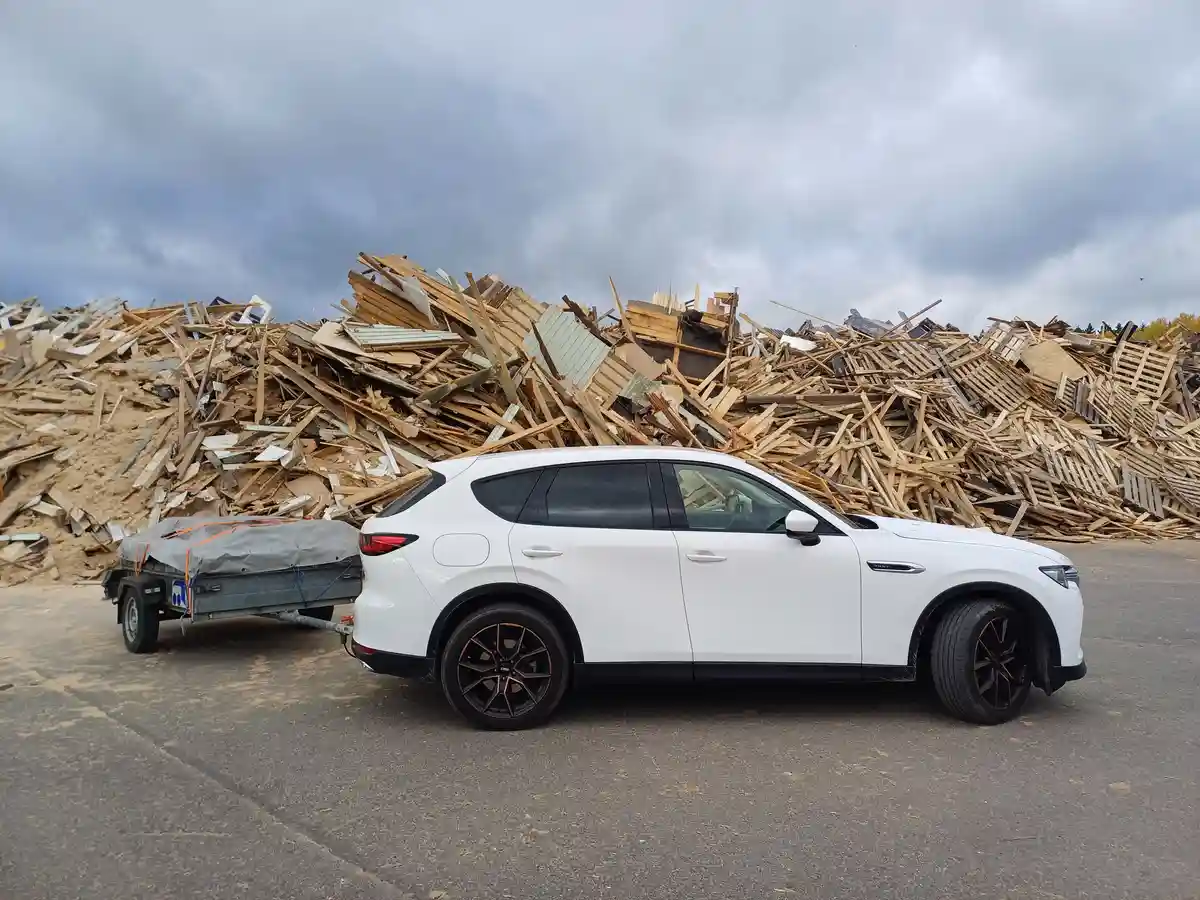 A white SUV with black wheels is parked on asphalt, towing a small trailer covered with a tarp. Behind it is a massive pile…
