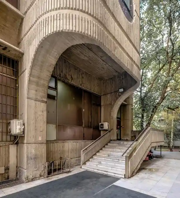 a photograph of a weathered, brutalist-style concrete building entrance with a large archway, stairs, and air conditioners…