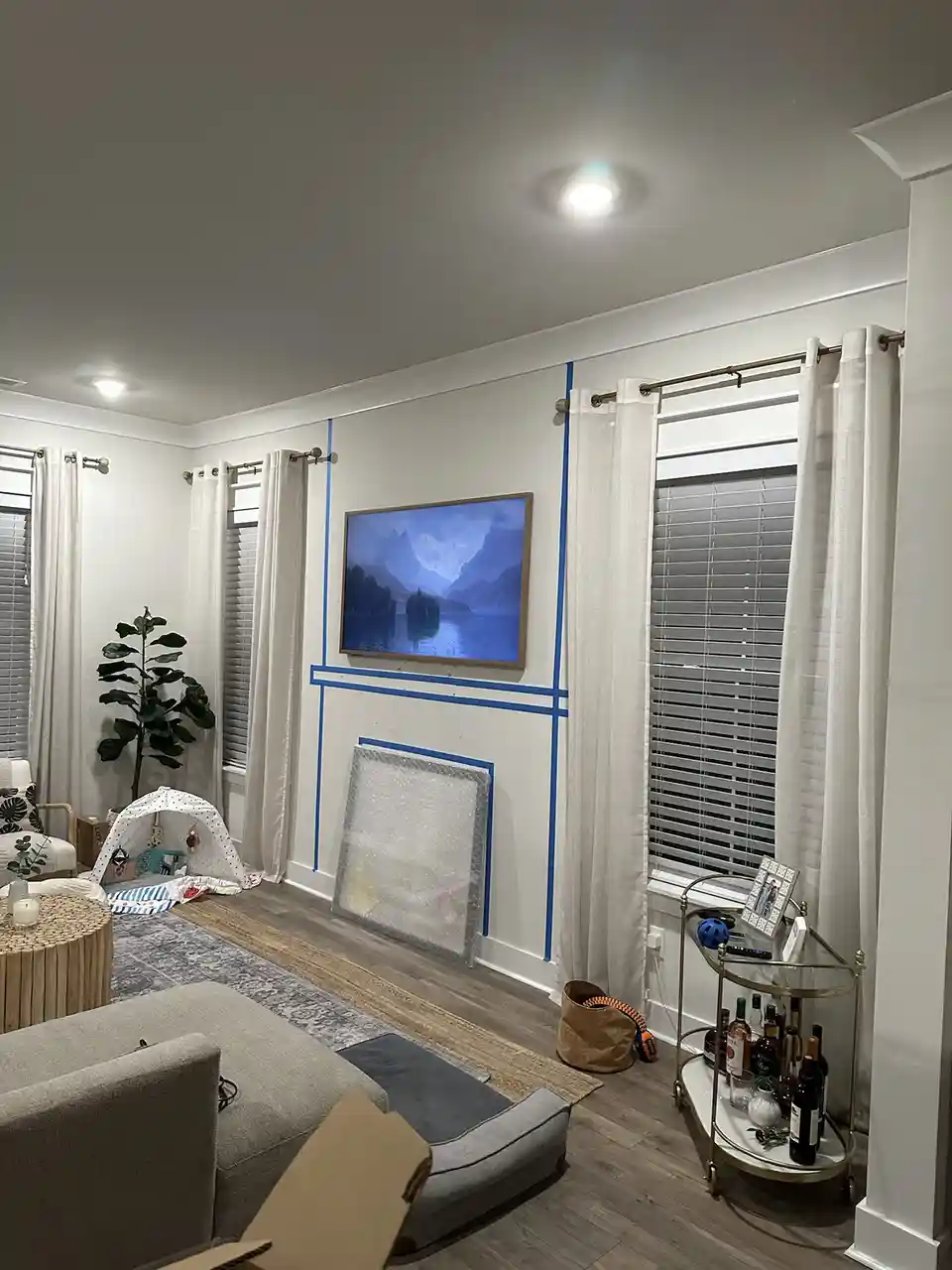 A living room with blue painter's tape outlining a rectangular wall space around a TV mounted on the wall, with a large…