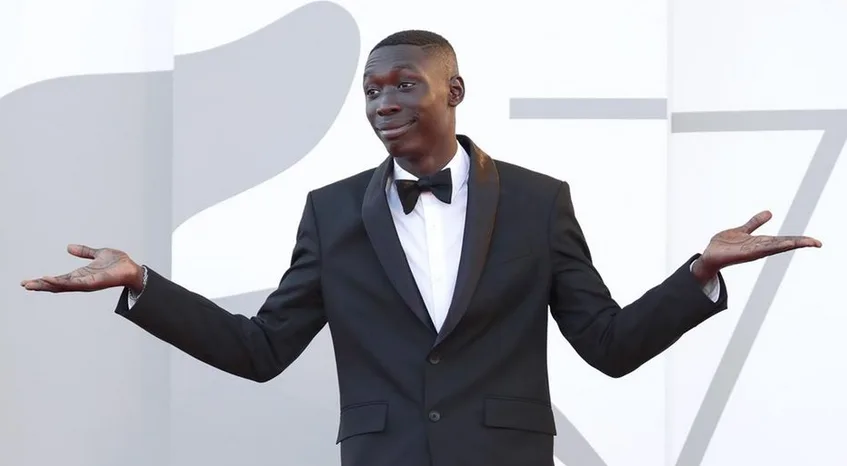 a man in a black tuxedo with a bow tie stands with arms spread wide, palms up, looking slightly amused or confused. Behind…