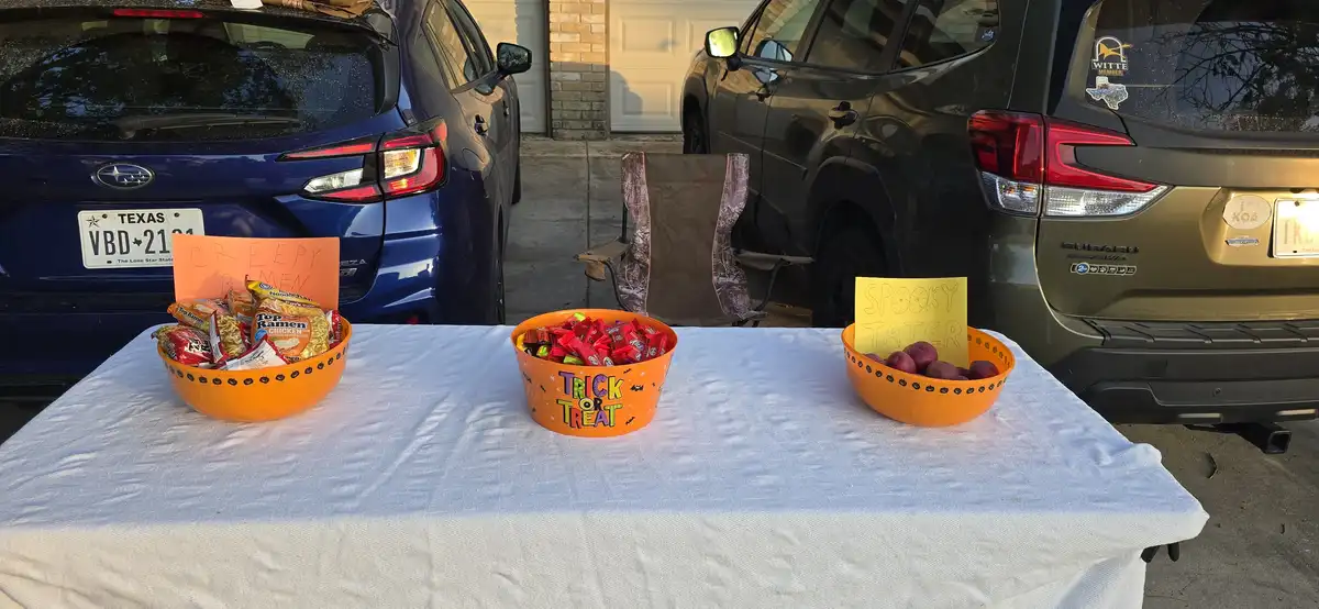 A white-clothed table outdoors holds three orange bowls: one with ramen noodles labeled 'creepy ramen,' one with candy…