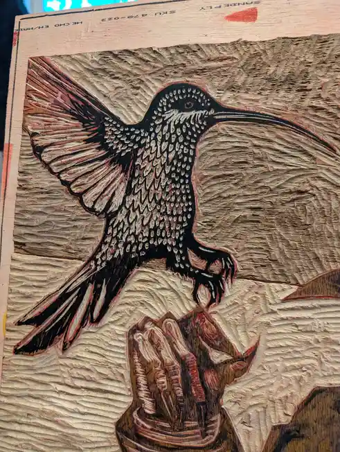 A close-up of a detailed wood carving depicting a hummingbird with outstretched wings perched on a flower, rendered with…