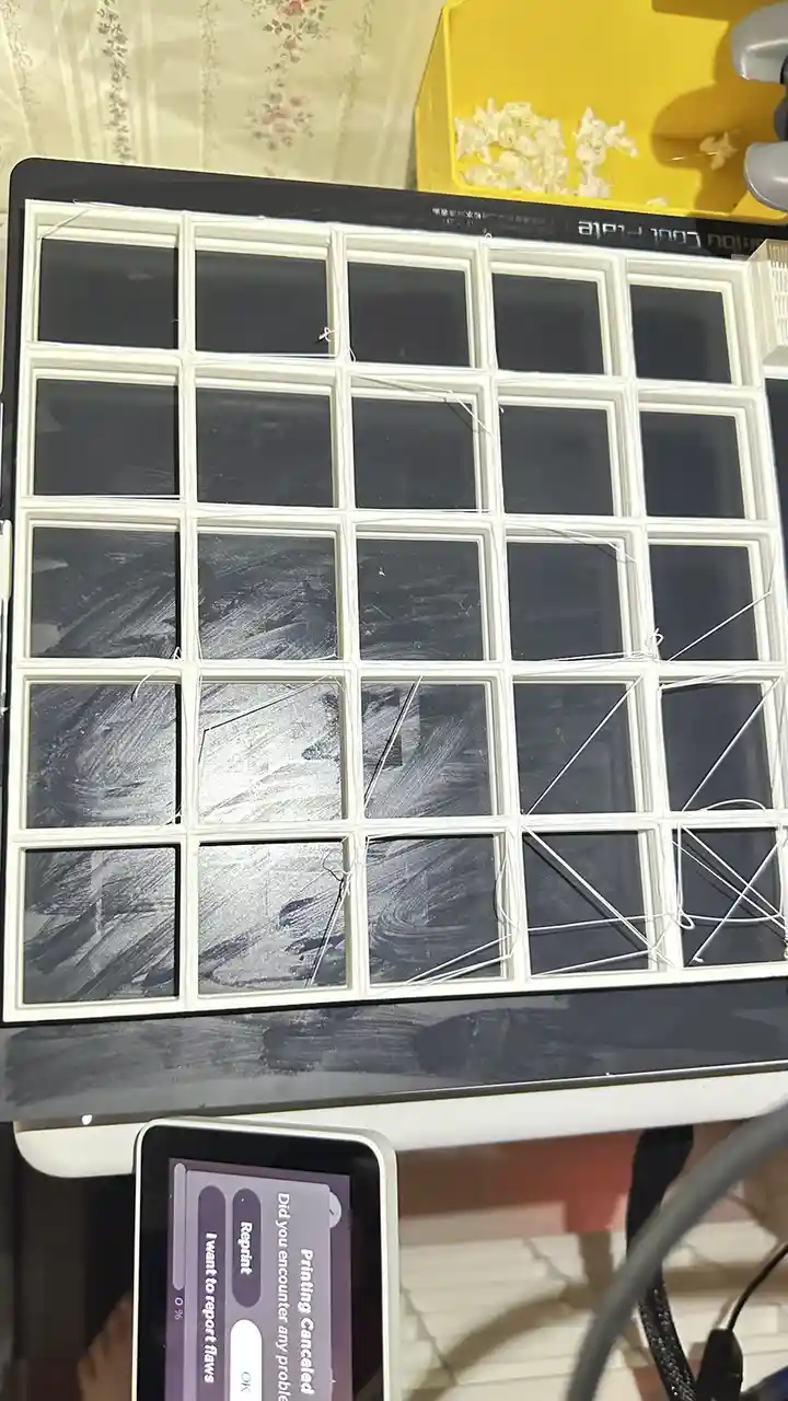 A 3D printer's build plate is covered in a grid of failed print attempts, with visible stringy filament and smudged…